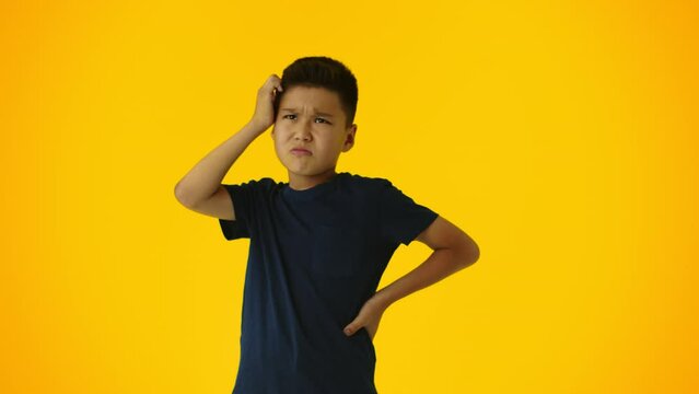 Young asian boy with a dissatisfied face scratching the back of his head thinking about what he wants to do on yellow background. Concept of lack of interesting activities for a child