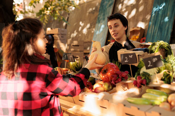 Female stall holder selling fresh organic bio products on stand, talking to customer about healthy eating and local natural veggies. Young woman asking small business owner about eco produce.