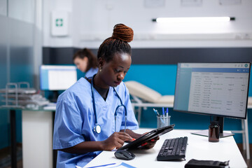 African american nurse checking appointments list on digital tablet in busy medical office. Adult...