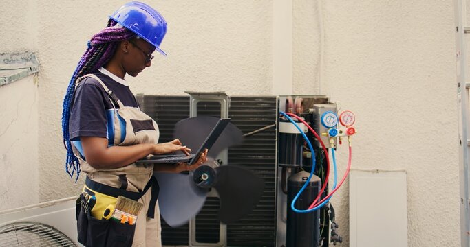 Adept african american repairman working on outside air conditioner holding laptop. Proficient worker optimizing HVAC system performance, ensuring it operates at maximum capacity