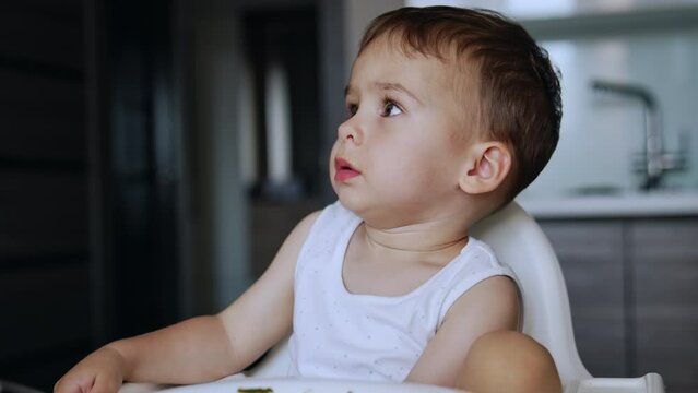 Cute toddler in white bodysuit sits at high chair. Mom gives him food on the spoon but stubborn kid refuses to eat. Close up.