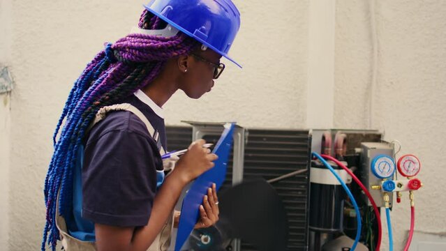 Skilled african american expert commissioned for outdoor air conditioner annual check up, writing findings on clipboard. Seasoned wireman doing hvac system investigation of damaged parts