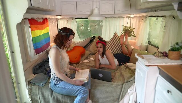 Two beautiful hippie girls relax on the bed inside the trailer, chatting, with a laptop and a book in their hands.