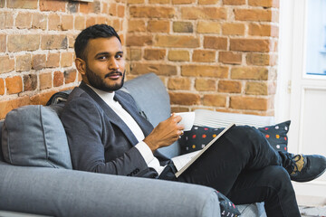 Indian man in a suit sitting on a sofa at a salon holding a cup of coffee. Reading while waiting. High quality photo