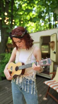 The beautiful hippie girl is singing and playing the guitar and singing near the trailer.