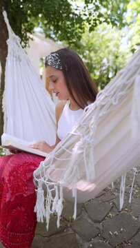 The beautiful hippie girl is sitting in a hammock on a summer day, reading a book.