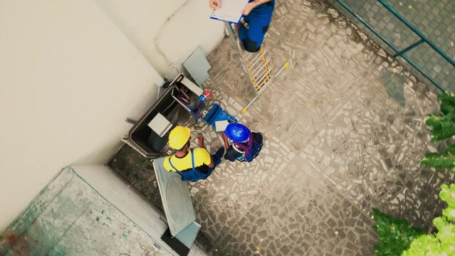 Drone shot of trained engineers cleaning and lubricating air conditioner components, verifying freon level and calibrating thermostats while coworker checks maintenance plan on clipboard