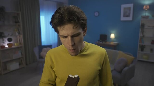 Young man eating ice cream, while standing in the living room close up. A man grabs his cheek, with a suffering expression, feels pain in his teeth. Home medicine concept. HDR BT2020 HLG Material.