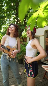 Two beautiful hippie girls are playing the guitar and laughing near the trailer.