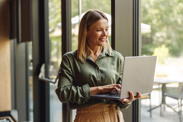 Smiling business woman working on laptop in cozy office standing near windows