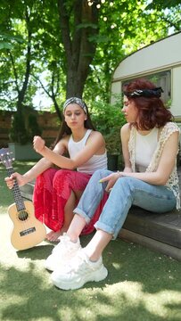 Two beautiful hippie girls are talking sitting on the terrace near the trailer with a guitar.
