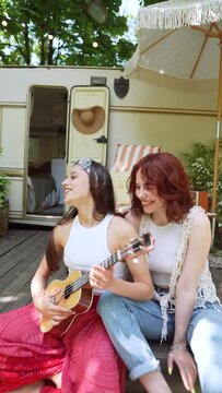 Two beautiful hippie girls are laughing and singing a song sitting on the terrace near the trailer with a guitar.