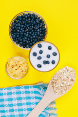 Drink in a glass on a yellow background with bowl of cornflakes and spoon with cereals. Glass of cold milkshake with fresh blueberries on top.