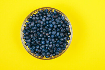 Glass bowl filled with ripe blueberries on a yellow background. Top view