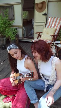 camera rolls on Two beautiful hippie girls are singing a song sitting on the terrace near the trailer with a guitar.