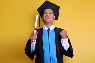 Excited man in graduation cap and ceremony robe clenching his fist and eyes closed celebrating...