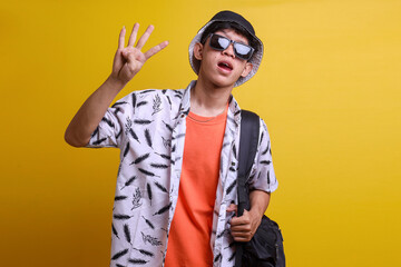 Happy young male tourist wearing sunglasses, bucket hat and backpack showing number four use finger standing against yellow background.