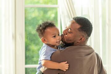 Happy african black parents dad father throw baby son on piggyback and neck riding teasing fun near window. Black baby son and daddy enjoy teasing kiss cheek and neck riding at window light