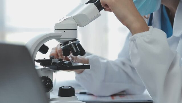 Medical worker in lab coat and sterile mask, doing a microscope analysis while her colleague are working behind