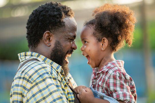 Happy african black parents dad father with daughter child on piggyback teasing fun in garden greenhouse. Black daughter kiss cheek dad and neck riding in vegetable greenhouse garden in light sunset