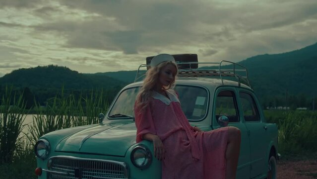 A girl in a vintage dress leans on a turquoise-colored retro car against a backdrop of mountains