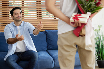 Fototapeta premium young gay couple looking at boyfriend, holding bouquet of roses and small gift box behind his back for surprise at home
