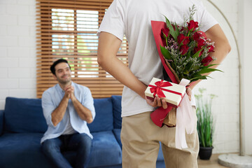 young gay couple looking at boyfriend, holding bouquet of roses and small gift box behind his back...