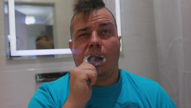 Drunk adult man brushing teeth after party with an unfocused look, relaxed face one eye closed, sleepy millennial guy in bright t-shirt going to work in big city.