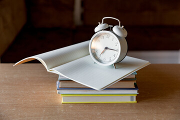 A stack of books, an open notebook and an alarm clock. Morning, the beginning of a new day of knowledge. The rising sun is reflected in the glass.