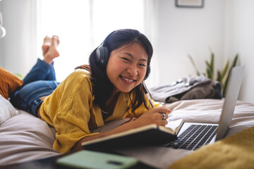 Happy asian teen student doing homework at home bedroom lying on bed looking at camera.Chinese teenage girl using laptop