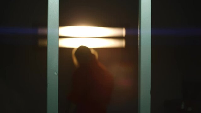 Female prisoner in orange uniform stands near barred window in prison cell. Terrified woman criminal shakes and looks at camera. Inmate serves imprisonment term in jail or detention center. Portrait.
