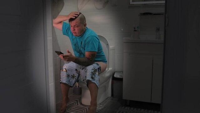 Caucasian man with constipation sits toilet with phone in hand, leaning against wall, continues to send messages and read the news.