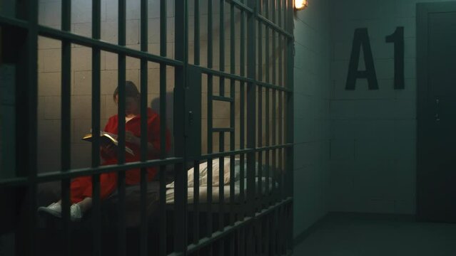 Female prisoner in orange uniform sits on the bed behind bars, reads book or Bible in prison cell. Woman criminal serves imprisonment term for crime in jail, detention center or correctional facility.