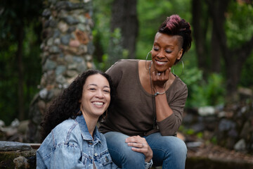 Sitting next to an old stone chimney, a multiracial lesbian couple shares a casual, romanic moment of everyday life. While one of them smiles to the camera, the other glances lovingly at her partner.