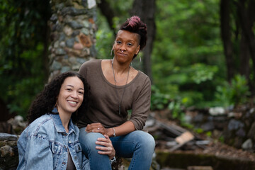 Two women poses for their engagement photos together. They are seated in a rustic, natural area. They are affectionately touching each other, reflecting their loving, nurturing relationship.