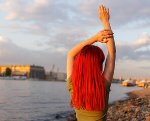 A young girl with red loose hair with her hands raised poses for the camera in the sunset rays on the river bank. Beautiful model posing.  Blurred background.