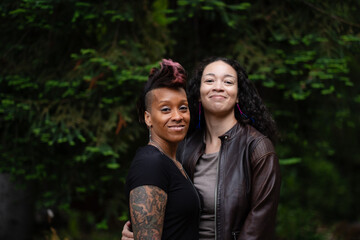 In front of lush greenery in a forest, two women stand close together with their arms around each other. They are posing four couples photos, celebrating their engagement.