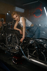 Naklejka na ściany i meble Beautiful woman mechanic posing for camera nearby motorcycle at workshop
