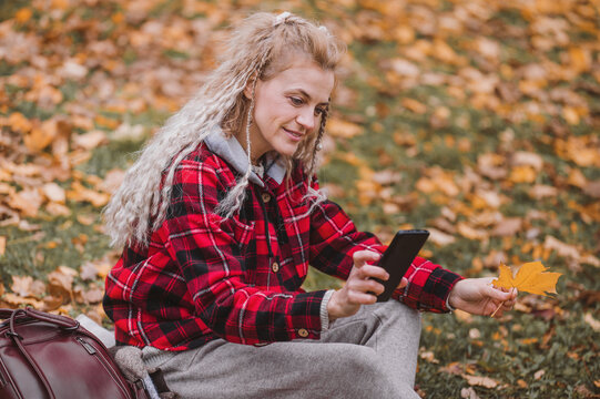 Middle aged woman shoot yellow leaves with smartphone. Mature blonde woman lies on a plaid and plays with yellow leaves in the autumn park