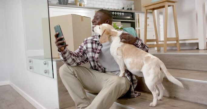 Happy african american man sitting on stairs using smartphone, with his pet dog at home, slow motion