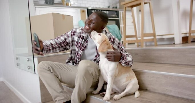 Happy african american man sitting on stairs taking selfie with his pet dog at home, slow motion