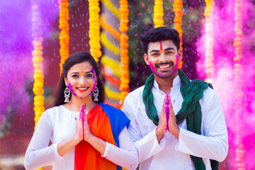 Happy young couple with color on face saying namaste againt flower decorated background - concept of holi festival greeting, wishes and Indian traditional culture
