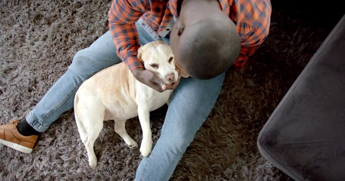 Happy african american man sitting on floor at home, petting his pet dog, slow motion