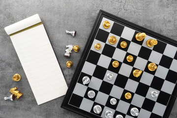 Chess board with chess pieces and notebook on stone texture background. Chess gold with silver battle. Top view.
