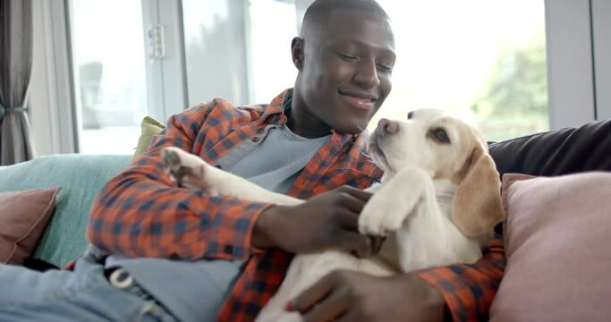 Happy african american man sitting on sofa, embracing his pet dog at home, slow motion