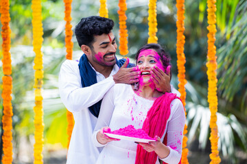 Obraz premium Happy indian young husband applying holi colors to wife face by holding holi powder plate during celebration holi festival - concept of fun, togetherness and colourful Indian tradition