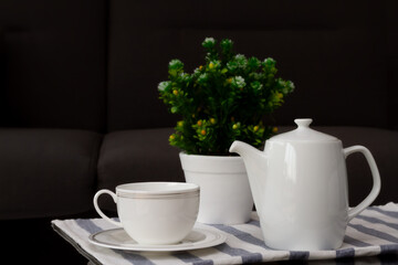 White coffee cup or teacup and jug with blurred flowerpot put on coffee table in the living room. Selective focus and copy space.