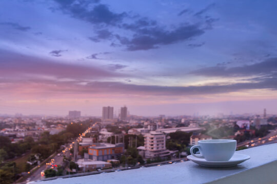 White coffee cup and softy smoke put on sky terrace border with high view blurred morning city scape and traffic on road. Selective focus and copy space
