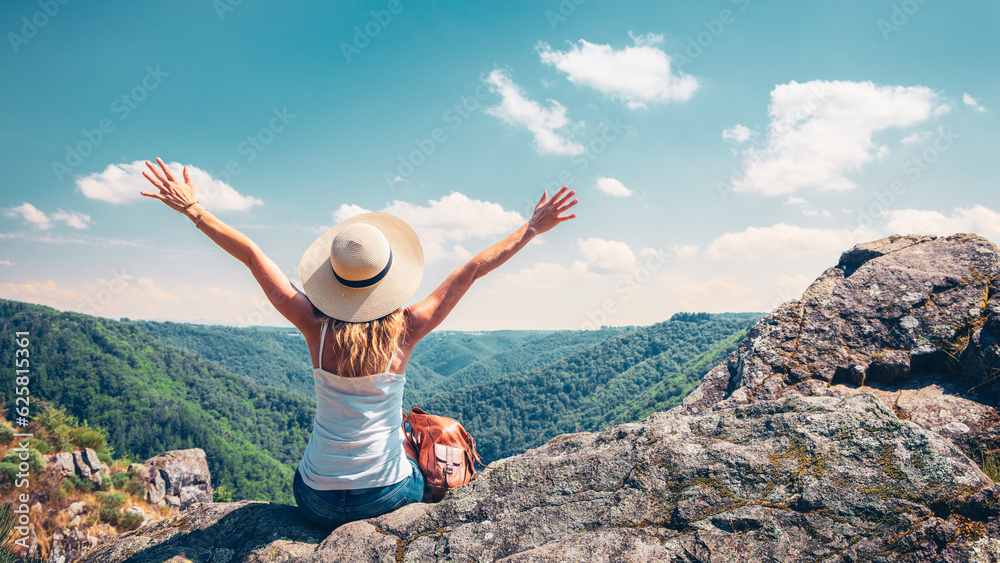 Wall mural woman with hat and backpack sitting on mountain peak enjoying beautiful view (dordogne in france)- a