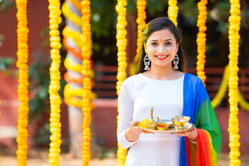Portrait shot of girl standing with pooja thali or plate against flower decoration background...